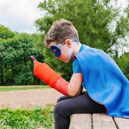 Junge mit Gipsarm und Superman Outfit schaut auf sein Smartphone